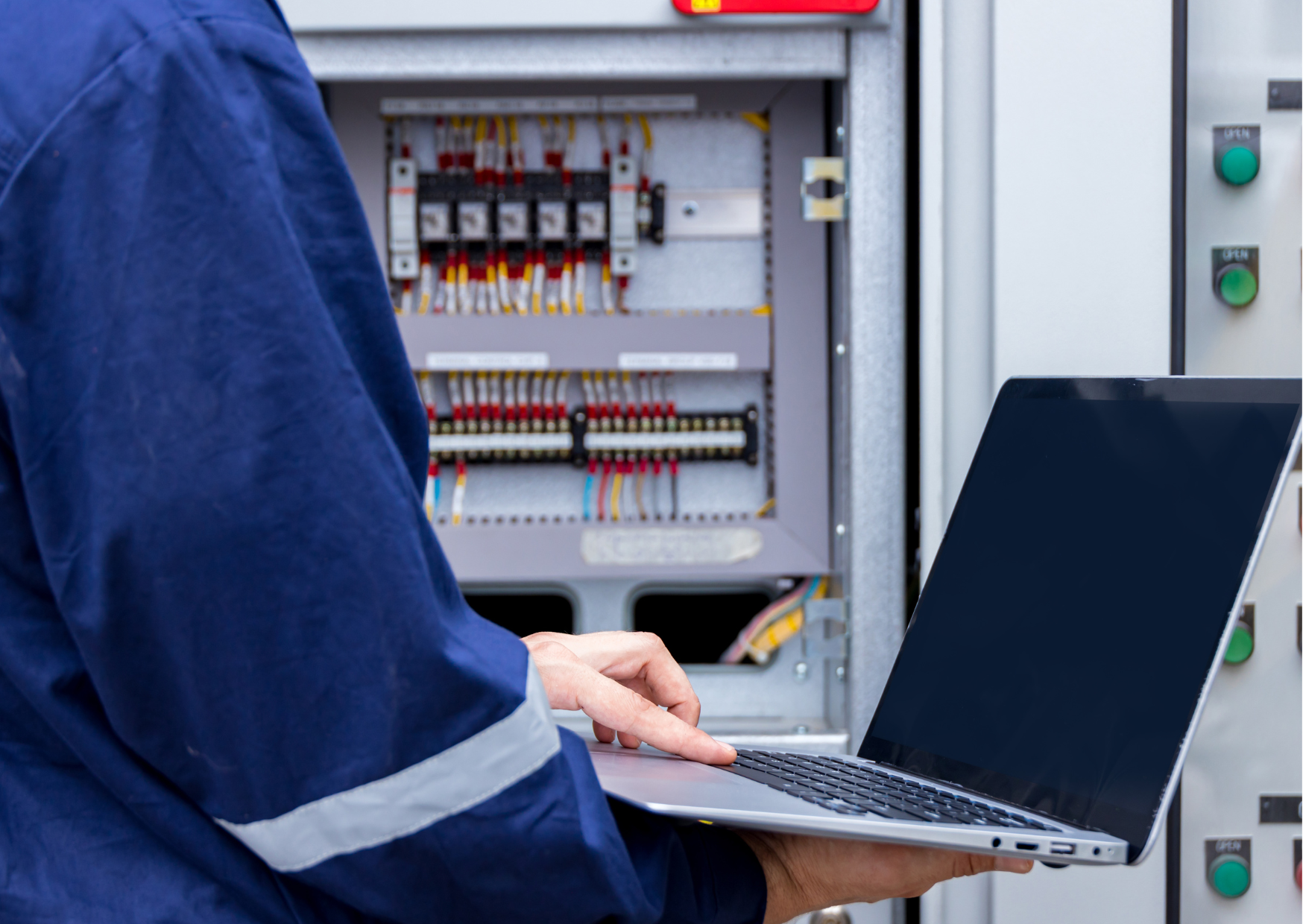 Un electricista con uniforme azul usa una computadora portátil frente a un panel de control eléctrico abierto con cables y disyuntores.