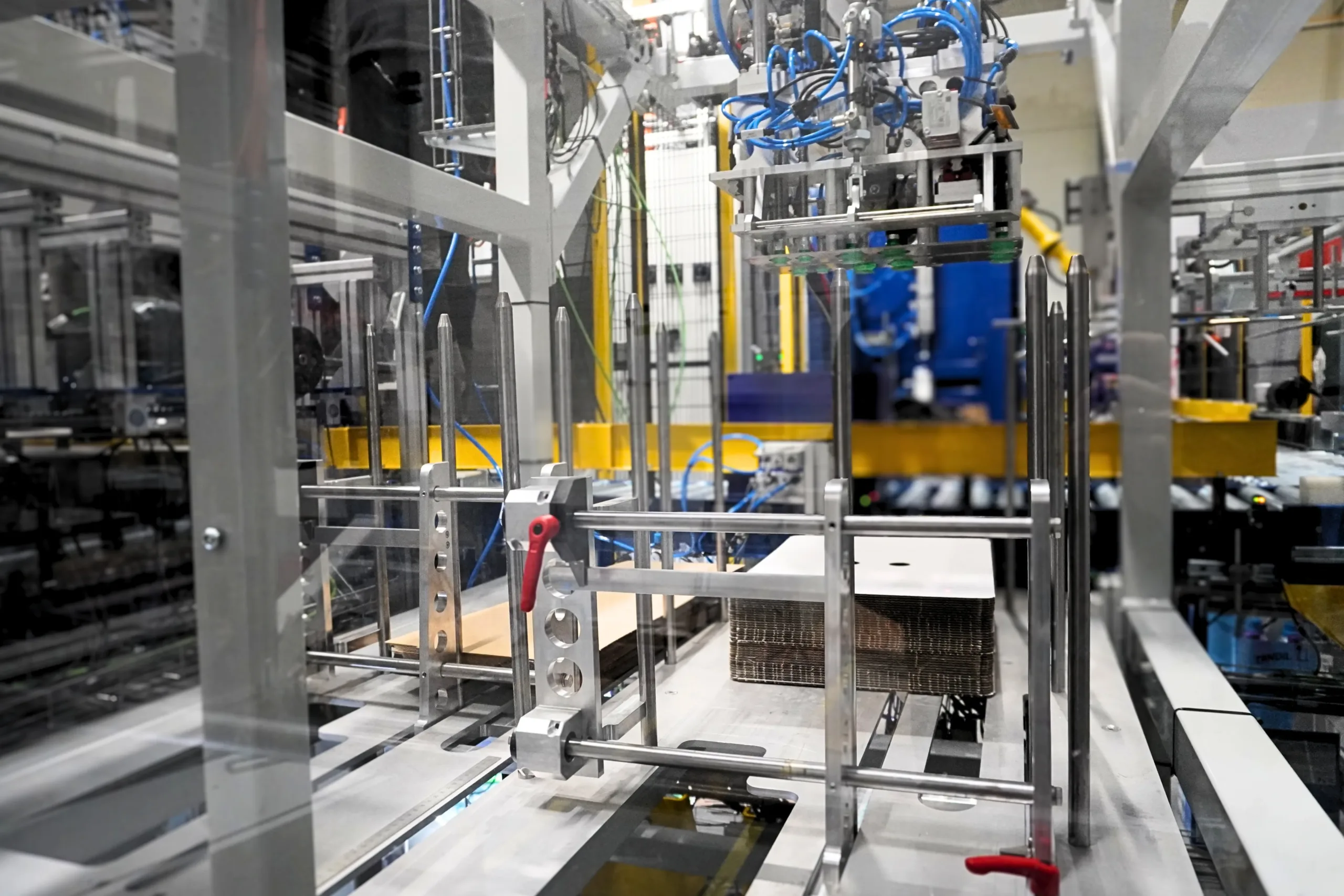 34972615 (7) Wnętrze automatycznej składarki i pakowarki kartonów. Z góry widoczna jest głowica chwytaka z niebieską instalacją pneumatyczną, a poniżej stos płaskich arkuszy tektury falistej w metalowej prowadnicy.